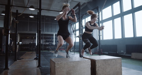 Two young happy beautiful athletic Caucasian women exercising together in large gym jumping on big...