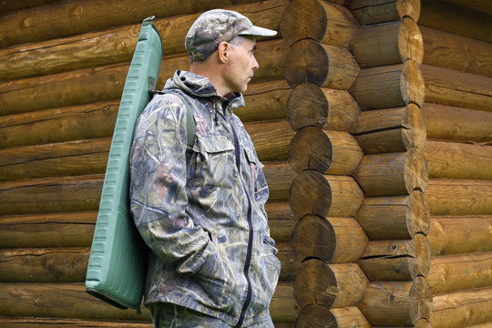 a hunter with a gun in a case on his shoulder stands near a log house