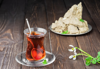 Armudu glass with red tea on a dark wooden table, next to a bouquet of spring flowers. A plate with halva with sesame seeds decorated with mint leaves Shallow depth of field.