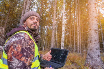 Ecologist works in the forest on the computer, shows a thumb. The gesture is OK. The forester is watching the development of forest stands. Forestry and afforestation.