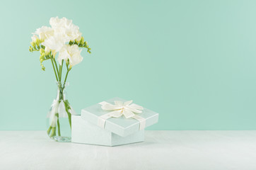 Festive background with opened square gift box and fresh white freesia flowers in elegant transparent vase on green mint menthe wall, white wood table.
