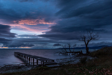 View of a pier and tree on a lake at dusk, beneath a dramatic, moody sky