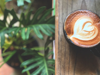 Latte coffee in black cup on vintage wooden table with natural light and Monstera deliciosa background  .