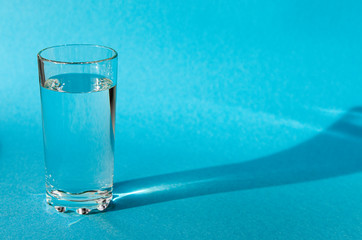 A glass with clean clear clear water stands on a blue background with green shadows. Clean water