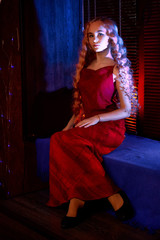 Portrait of young elegant tender blonde teenage girl in red dress on the window sill in dark room with blue light. Caucasian female model with very long hair posing indoors