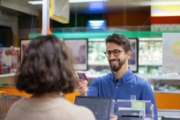 Smiling man giving credit card to cashier. Customer talking with cahier while buying goods in...