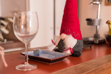 Christmas female elf sitting on a table ready to have dinner and wine. Beautiful original stuffed toy close up. Has a high red pointed hat hiding its eyes and long braid hair. Ready for december 25.