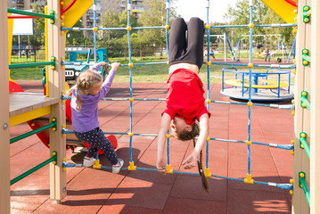 Two happy caucasian girls having fun on playground in summer, climbing the rope net. The older girl is hanging upside down - Powered by Adobe