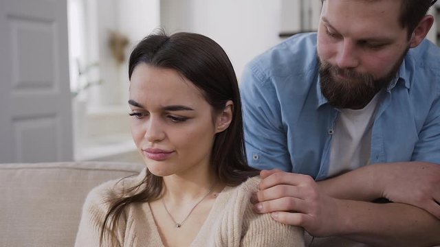 Handsome bearded young man trying to calm his upset hapless girlfriend and hugging her the back