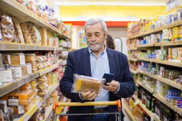 Man holding smartphone and choosing products in store. Focused bearded mature man holding mobile phone and choosing goods in grocery store. Shopping and technology concept