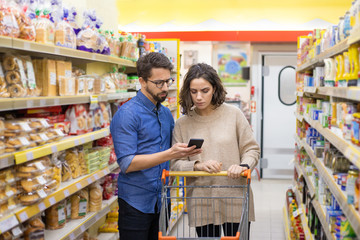 Couple using smartphone in grocery store. Focused young man and woman standing with shopping trolley, holding mobile phone and buying products in supermarket. Shopping concept