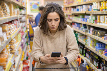 Woman leaning at cart and using smartphone in store. Young woman reading checklist via smartphone and choosing products in grocery store. Supermarket concept