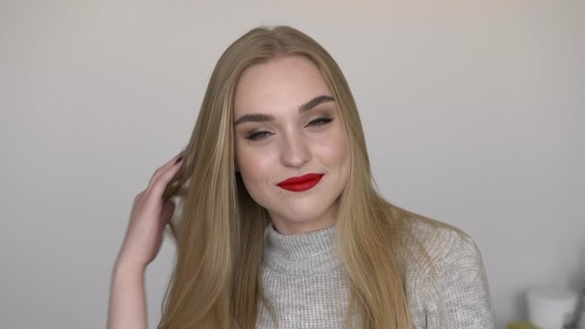Indoors slow motion closeup face shot of young smiling woman smoothing her long blond hair by turning head, bright red lips, dressed grey sweater, posing against white wall natural light