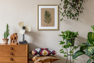 Stylish retro home staging of living room with gold mock up poster frame, design furnitures, a lot of plants, pillows, retro fan and elegant personal accessories. Vintage home decor. Template. 