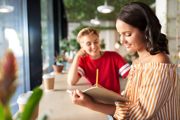 people and friendship concept - young woman with friend at cafe writing to notebook