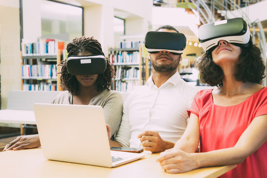 Multiethnic team of adult students enjoying VR experience together. Man and women in virtual reality glasses, sitting at desk with laptop, enjoying experience. Group VR experience concept