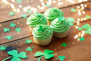 st patricks day, food and holidays concept - green cupcakes and shamrock on wooden table over festive lights