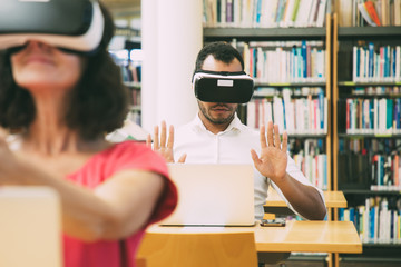 Students using VR simulators for studying. Man and woman in virtual reality headsets sitting at desks with laptops and touching air. VR technology for education concept