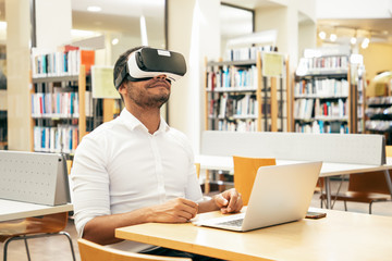 Happy adult male student enjoying VR experience in library. Man wearing virtual reality glasses, sitting at desk with laptop and smiling. Virtual video concept