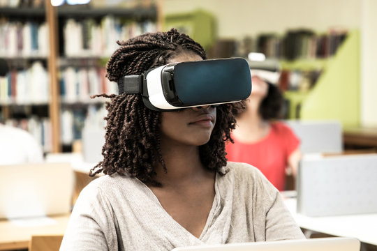 Interracial group of students using VR experience for work in library. Young black woman wearing virtual reality headset, sitting at desks, using laptop. Innovation technology concept