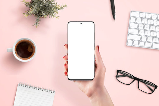 Phone mockup in woman hand. Clean scene for app promotion. Top view, flat lay. Pink work desk in background with coffee, keyboard, plant, glasses, pad and pen