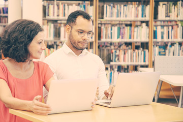 Positive couple of adult students doing and discussing research in library. Man and woman in casual sitting at desk, using laptops, typing, talking. Apprenticeship concept