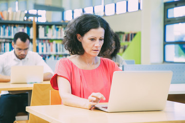 Shocked female adult student taking online test in classroom. Serious Latin man sitting at desk and using laptop. Bookshelves in background. Education concept
