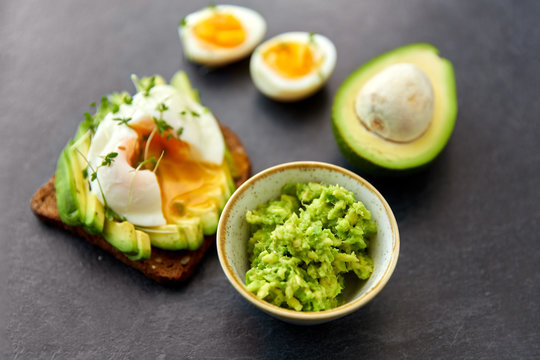 food, eating and breakfast concept - toast bread with sliced avocado, pouched egg and greens on slate stone table