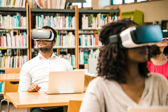Diverse group of students watching virtual video tutorial in library. Man and women wearing virtual reality headsets, sitting at desks with laptops. Hardware for library concept