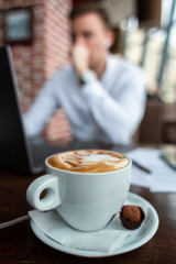 White cup of coffee and a sweet on a saucer on the table at a cafe. Young businessman with a laptop on the background. Freelance and selfeployement concept.