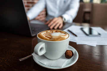 White cup of coffee and a sweet on a saucer on the table at a cafe. Young businessman with a laptop on the background. Freelance and selfeployement concept.