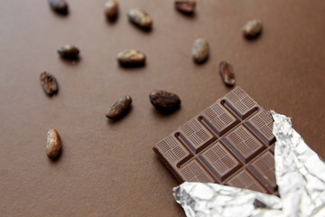 sweets, confectionery and food concept - dark chocolate bar in foil wrapper and cocoa beans on brown background