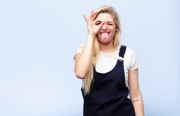 young pretty blonde woman smiling happily with funny face, joking and looking through peephole,...
