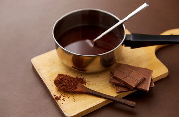 sweets, confectionery and culinary concept - pot with melted hot chocolate, cocoa powder in spoon and wooden board on brown background