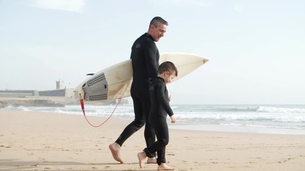 Father and son in wetsuits walking on beach. Side view of happy father holding surfboard and walking with cute little son on sandy beach, tracking shot. Surfing concept
