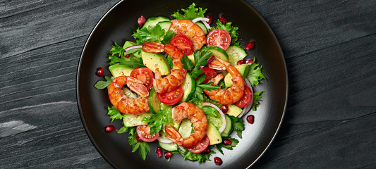 Shrimps salad, avocado, lettuce, cherry tomatoes. Home made vegetarian shrimp salad. Seafood concept. Tasty veg mixed leaves, grilled prawn shrimps in plate on black wood background.