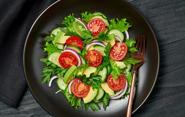Fresh salad with cherry tomatoes, avocado,cucumeber and lettuce on dish. Healthy vegetarian meal concept. Tasty mixed leaves, tomatoes and mix vegetable salad on black wood, top view.