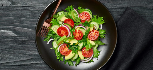 Fresh salad with cherry tomatoes, avocado,cucumeber and lettuce on dish. Healthy vegetarian meal concept. Tasty mixed leaves, tomatoes and mix vegetable salad on black wood, top view.