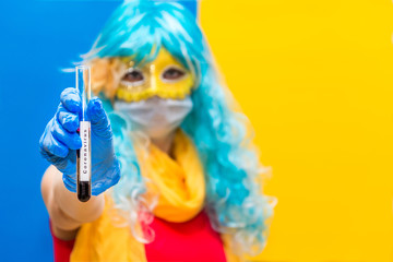 Concept of protection against coronavirus at the carnival venice. A bright woman in a blue wig, yellow mask and medical gloves holds a test tube with a blood test for Virus crown. Copy space