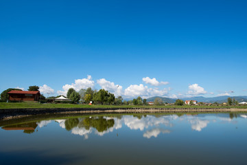 Fototapeta premium Paesaggio con lago di campagna e riflesso del cielo e delle nuvole sull'acqua con case e alberi