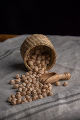 Close-up of basket lying with raw chickpeas on gray linen cloth, wooden table, black background and dark light in vertical