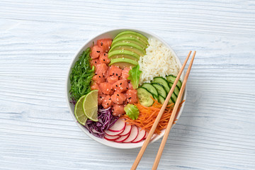 Hawaii salmon poke, rice, seaweed, tomato, cucumber, radish, carrot salad bowl. Home made vegetarian poke. Seafood tasty diet concept. Asian salmon poke bowl on white wood background.