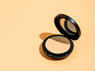 Compact powder on yellow or cream background. Female pressed powder in ajar opened black plastic case with mirror, copy space for text or design. Hard light