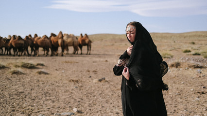 beautiful woman national eastern clothes black color,veil,hijab,abaya ornament her head,portrait steppes camels,strong wind,desert,wildlife,well,decoration,caucasian,slow ,sun