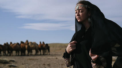 beautiful woman national eastern clothes black color,veil,hijab,abaya ornament her head,portrait steppes camels,strong wind,desert,wildlife,well,decoration,caucasian,slow ,sun