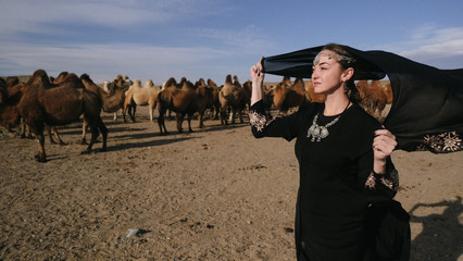 beautiful woman national eastern clothes black color,veil,hijab,abaya ornament her head,portrait steppes camels,strong wind,desert,wildlife,well,decoration,caucasian,slow ,sun