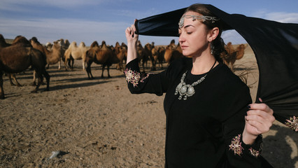 beautiful woman national eastern clothes black color,veil,hijab,abaya ornament her head,portrait steppes camels,strong wind,desert,wildlife,well,decoration,caucasian,slow ,sun