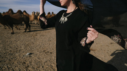 beautiful woman national eastern clothes black color,veil,hijab,abaya ornament her head,portrait steppes camels,strong wind,desert,wildlife,well,decoration,caucasian,slow ,sun