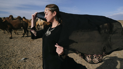 beautiful woman national eastern clothes black color,veil,hijab,abaya ornament her head,portrait steppes camels,strong wind,desert,wildlife,well,decoration,caucasian,slow ,sun