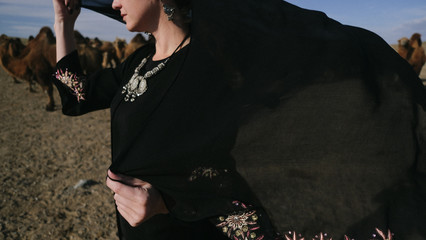 beautiful woman national eastern clothes black color,veil,hijab,abaya ornament her head,portrait steppes camels,strong wind,desert,wildlife,well,decoration,caucasian,slow ,sun